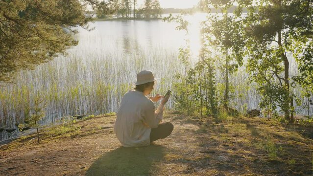 Young man surfs internet on smartphone sitting on river bank. Guy rests near water with bulrush and rocks at sunset. Tree branches sway in light wind