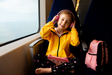 A girl on a train listens to music in headphones with a gadget in her hands. The girl is 6 years old. She goes on vacation with her parents. Family vacation. Gadgets. Vacation. Summer.