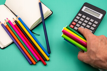 Top view of a hand pressing buttons on a calculator with a handful of colored markers in his hand and a green table with a notebook, colored pens and a calculator. Back to school concept. 