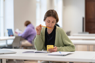 Focused pensive middle-aged woman studying in library holding phone, learning online, using mobile...