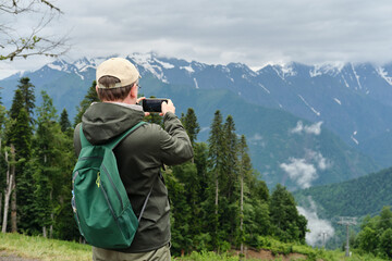 Back view of middle-aged man with backpack taking a photo of the mountains on his smartphone. 