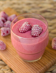 A glass of raspberry smoothie and two frozen raspberries. A glass of cooling drink on a wooden board with raspberry berries