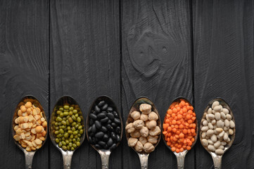 Variety of legumes in old silver spoons on a black wooden background.