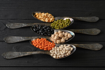 Variety of legumes in old silver spoons on a black wooden background.