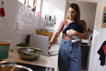 Caucasian mid adult mother using laptop while carrying newborn baby in kitchen at home