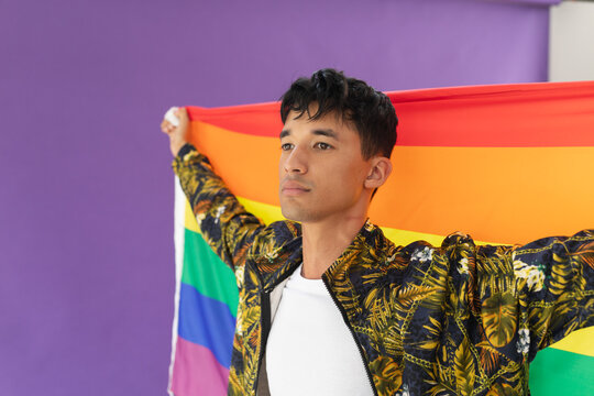 Biracial gay man holding lgbt flag around his back against purple background