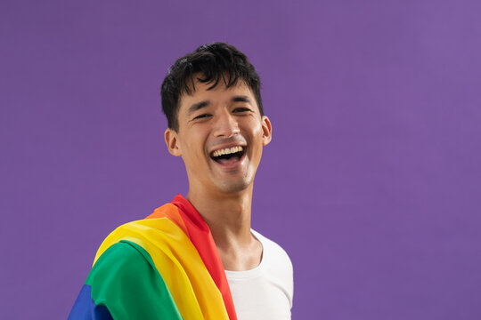 Portrait of biracial gay man with lgbt flag on his shoulder smiling against purple background