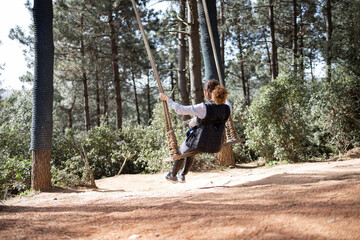 Fototapeta premium Young adult woman in rustic swing at outdoor. Young girl traveler sitting on the swing in beautiful nature place among trees. Freedom concept. 