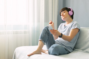 An adult woman in pink headphones sits on the sofa at home and listens to music