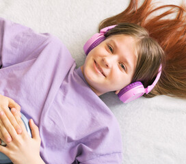 Happy teenage girl in lilac t-shirt lies on bed in pink headphones listens to music on CD player