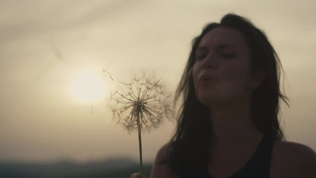 Slow motion video of a girl blowing a dandelion in the mountains at sunset. Woman rejoices chk child, happy adult in the mountains.