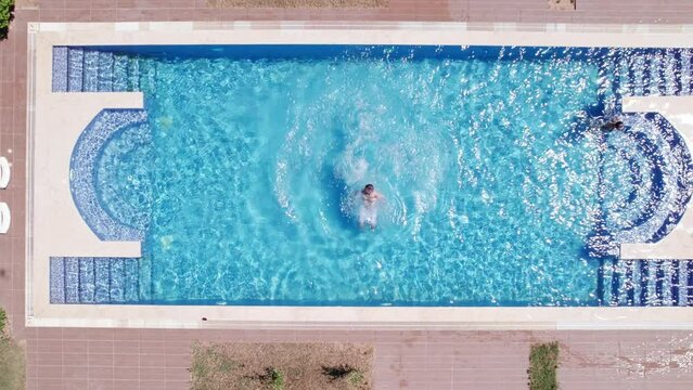Man doing a backflip dive stunt. Man dives to swimming pool. Real time top down aerial video footage of man jumping to the turquoise colored water of swimming pool. Summer leisure activities