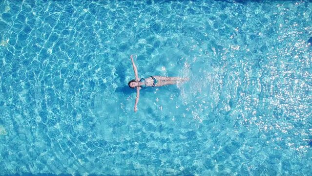 Top down aerial view of young woman relaxes lying backstroke on a rippling water surface in a swimming pool. Woman swims on a back. High angle view