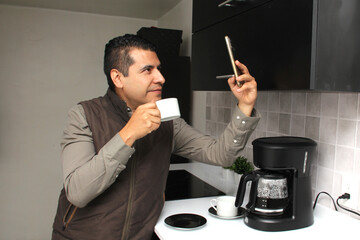 Latin adult man drinks freshly brewed coffee in the coffee maker in the kitchen of his home or office while using his cell phone to send messages, make calls at the beginning of the day

