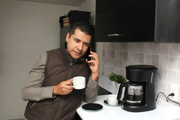Latin adult man drinks freshly brewed coffee in the coffee maker in the kitchen of his home or office while using his cell phone to send messages, make calls at the beginning of the day

