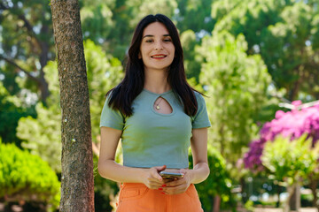 Young woman smiling confident wearing turquoise tee and orange short on city park, outdoors holding phone in hand and posing smiling for camera, self-confident