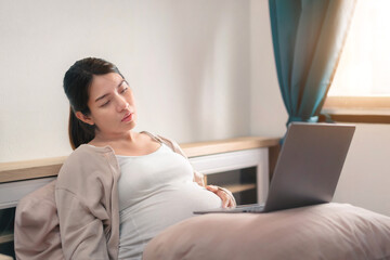 tired asian pregnant woman  and using laptop on bed for working at her bedroom, remote working, Online shopping, Women pregnancy concept, New normal and work from home concept.
