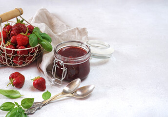 Strawberry jam with basil in the glass jar and a basket with berries at white table. Copy space
