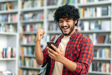 Gadgets concept, wireless technology. Cheerful indian or arabian guy, freelancer or student, using his smartphone, looks at a screen, rejoices in success, victory, makes yes gesture, smiling happily