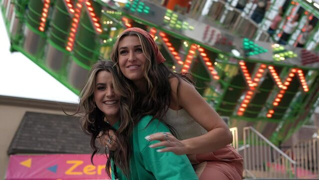 Two young women posing and waving while one rides the others back in front of ride at a carnival as it spins.