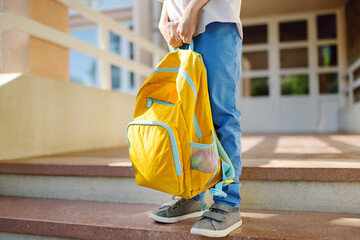 Little student with a backpack on the steps of the stairs of school building. Close-up of child legs, hands and schoolbag of boy standing on staircase of schoolhouse.Back to school concept. - obrazy, fototapety, plakaty