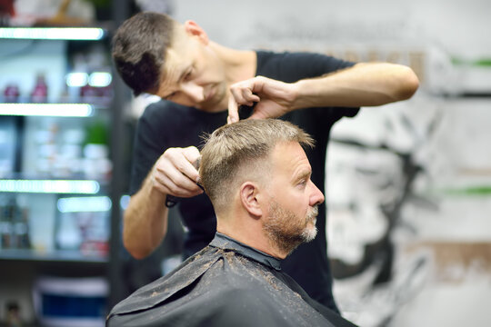 Hairdresser is cutting hair of handsome bearded mature man in salon. Stylist making hairstyle with electric shaver for person in barbershop. Services of a professional stylist. Fashion haircut