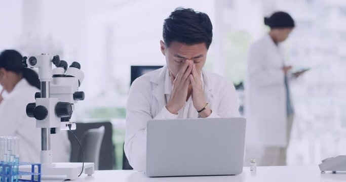 Stressed male scientist working on laptop in a lab. Young research biologist with a bad headache feeling tired from sitting at desk all day. Biotechnology chemist unhappy with statistics results