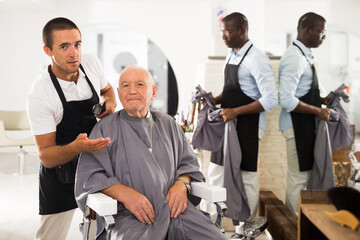 Fototapeta na wymiar Smiling gray-haired man sitting in hairdressing chair, discussing haircut with young professional barber