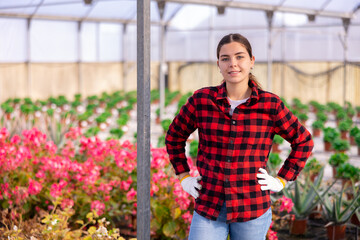 Fototapeta na wymiar Portrait of positive young female florist posing at her greenhouse on background with plantation of flowers