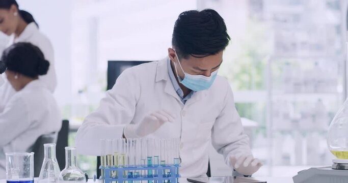 Young male biologist typing test tube results into a digital tablet in a COVID research lab. Scientist or chemistry specialist with a face mask, examing liquid in chemical glassware in clinical trial
