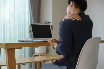 A young man sits at his desk at home using a computer. He puts his hands on the nape of his neck and shoulders when he feels pain in his muscles from sitting for a long time. - obrazy, fototapety, plakaty