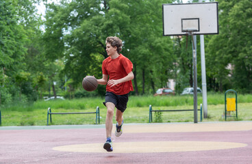 Fototapeta premium Teenager running in the stadium. Cute young teenager in red t shirt with a ball plays basketball on court. Sports, hobby, active lifestyle for boys 