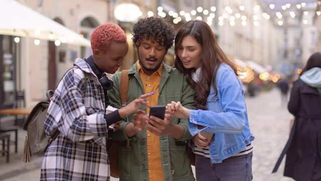 Happy young mixed-race females standing outdoor with male friend typing on smartphone and chatting choosing place to go Decorated street with restaurants on background Travelers concept, portrait shot