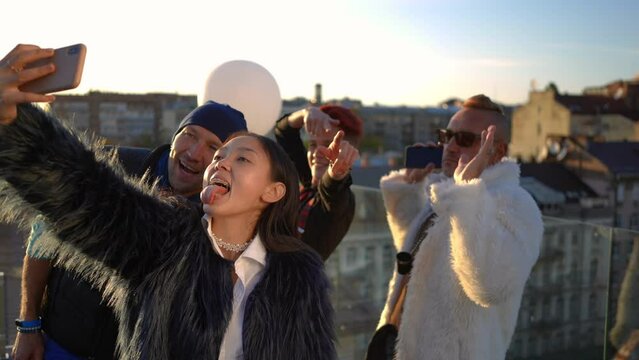 Asian woman taking selfie with Caucasian friends at sunset on rooftop making faces laughing. Positive relaxed confident group of people having fun grimacing photographing in sunshine in urban city