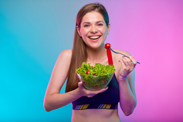 Smiling woman in sportswear eating green salad in glass bowl. Female fitness portrait isolated on neon multicolor background.