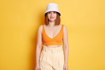 Portrait of beautiful delighted woman wearing top and panama standing against yellow wall, looking at camera with calm and optimistic facial expression.