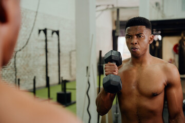 Naklejka na ściany i meble African American male holding weights during an intense workout. Male athlete looking at himself in the gym mirror while lifting dumbbells. High quality photo