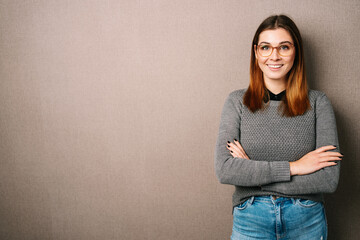 Young woman with glasses and folded arms looks at camera, brown wall with copy space