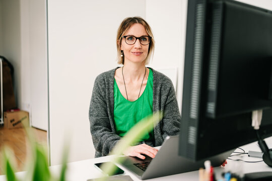 Young businesswoman with glasses sits at desk in office and looks at computer monitor