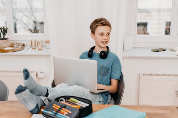 Schoolboy sitting relaxed at table doing homework with laptop