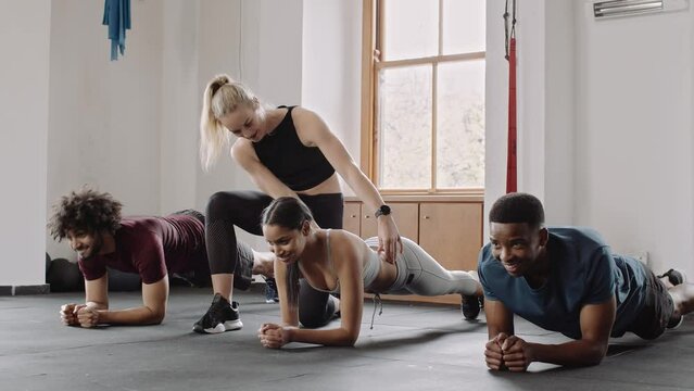 Caucasian trainer correcting client's form. Diverse group of individuals planking in group exercise class at an indoor fitness gym.