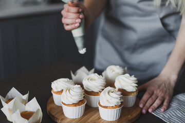 Young lovely woman pastry chef decorates cupcakes with whipped cream at home