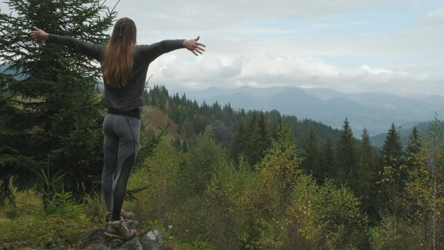 Long-red-haired young woman, standing on peak overlooking mountains and green forest, throws her arms out to the side from beauty nature. Cloudy, back view, copy space