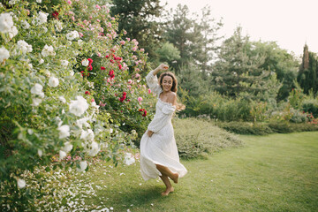 Obraz premium Young beautiful woman with long curly hair wearing white dress relaxing in blooming rose garden.