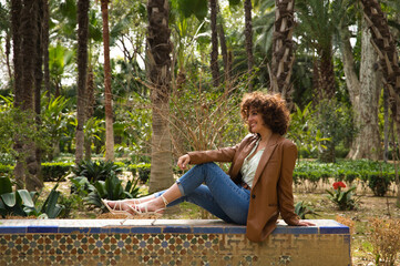 Beautiful woman with curly hair sitting on a bench in a park with large trees in a European city. The woman is dressed in modern and trendy clothes. Travel and holiday concept, rest and relaxation.