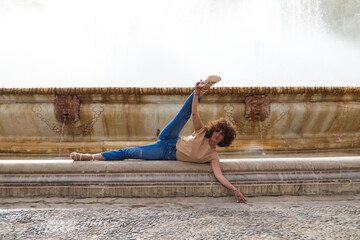 Naklejka premium Beautiful young woman who is a dancer is standing with her legs open in a large fountain in a square. The woman is athletic and has flexibility. Well-being and health concept.