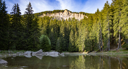 Lake with clear water and stone shore in spruce forest with fir trees against a daytime sky