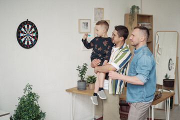 Little boy throwing darts in dartboard during game with his gay parents at home