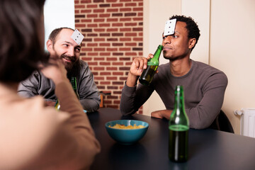 African american man playing guessing game with cards while sipping beer with friends at home. Happy smiling multiracial friends sitting at table in living room while enjoying fun leisure activity.