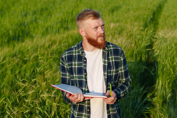 Naklejka na ściany i meble Portrait of an agronomist with a notebook in a wheat field. A farmer with a beard and a shirt prepares the field for the future harvest